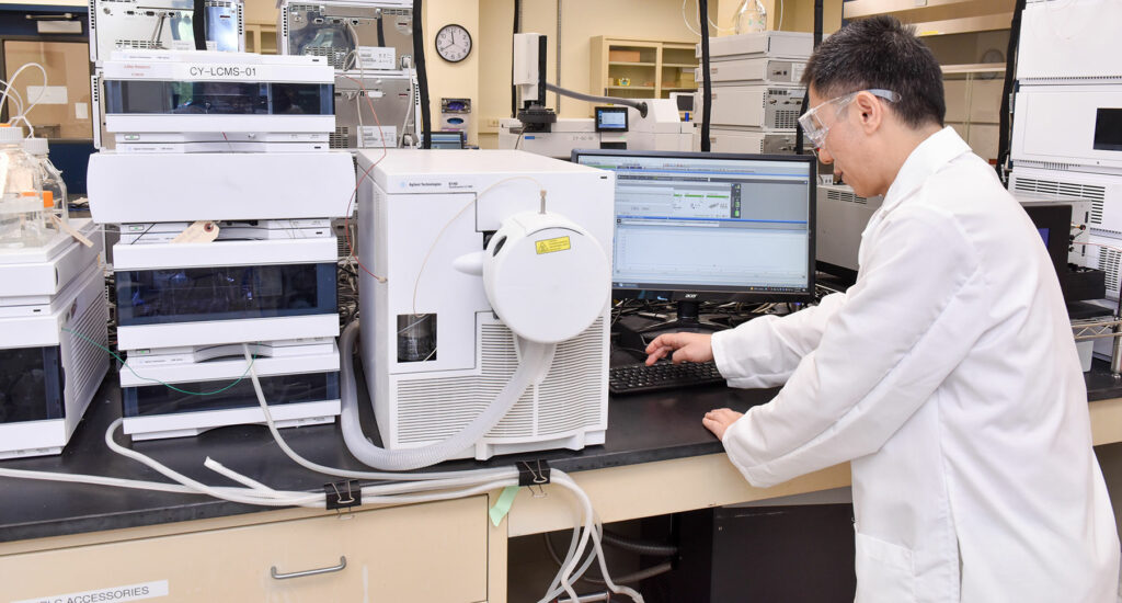 A scientist in a lab coat and safety goggles works at a computer connected to scientific equipment, possibly for chromatography analysis, in a laboratory setting.