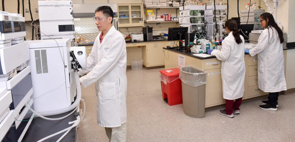 Three scientists in lab coats work in a laboratory. One operates equipment, while two others conduct experiments at a bench. The lab is equipped with various machines and storage cabinets.