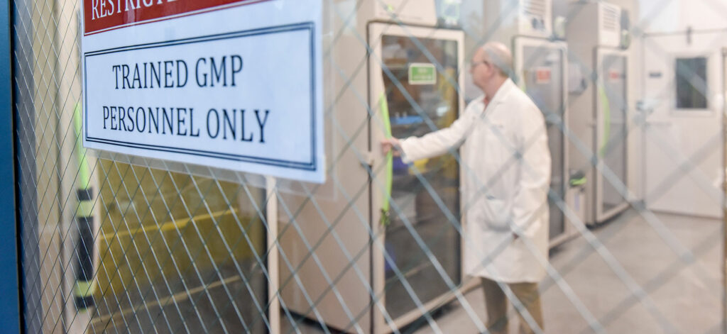 Person in lab coat entering a restricted area labeled for trained GMP personnel only, with equipment visible in the background.