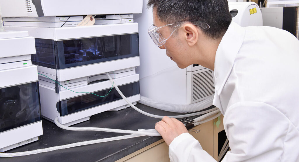 A person in a lab coat and safety goggles examines scientific equipment with tubes on a laboratory bench.