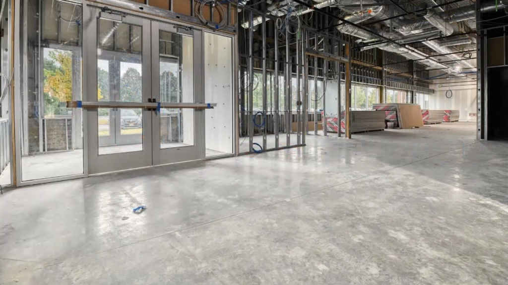 The interior of the unfinished commercial building reveals a state-of-the-art design with exposed framework, concrete floors, and sleek glass entry doors. Construction materials are visible in the background, hinting at its future as a cutting-edge facility.