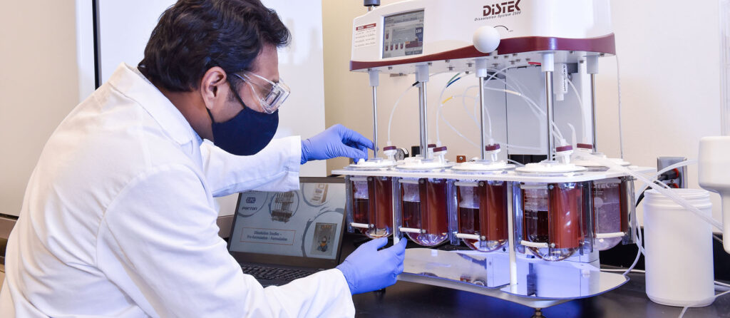 A scientist in a lab coat and mask conducts an experiment using a dissolution tester with multiple beakers, while checking data on a laptop.