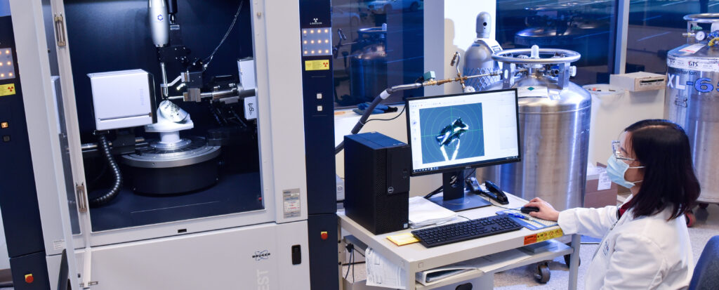 A researcher in a lab coat operates a computer next to a large analytical machine. The monitor displays data.