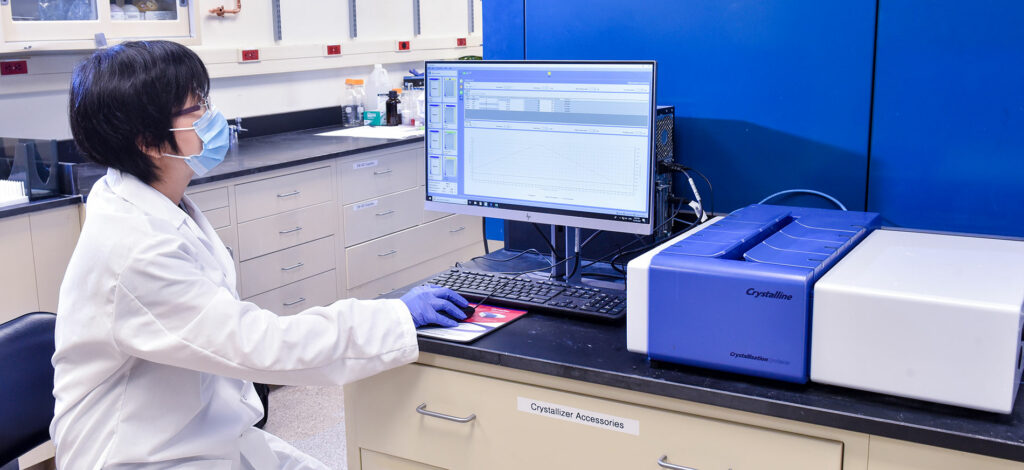 A person in a lab coat and mask sits at a desk, using a computer connected to a blue crystallization machine, in a laboratory setting.