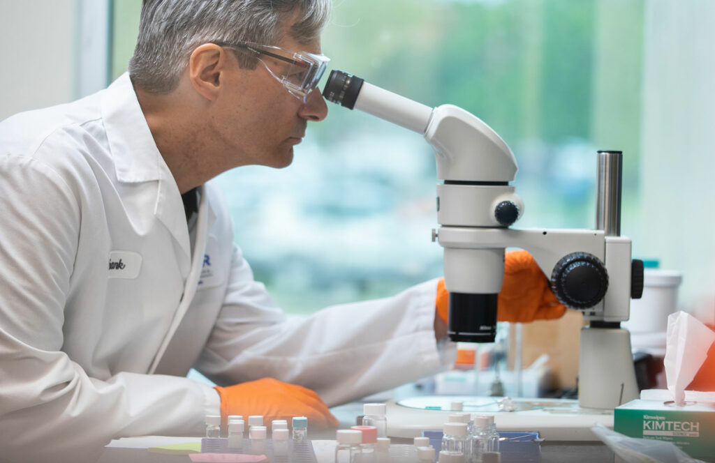 A scientist wearing a lab coat and safety goggles looks through a microscope in a laboratory, with orange gloves and vials on the desk.