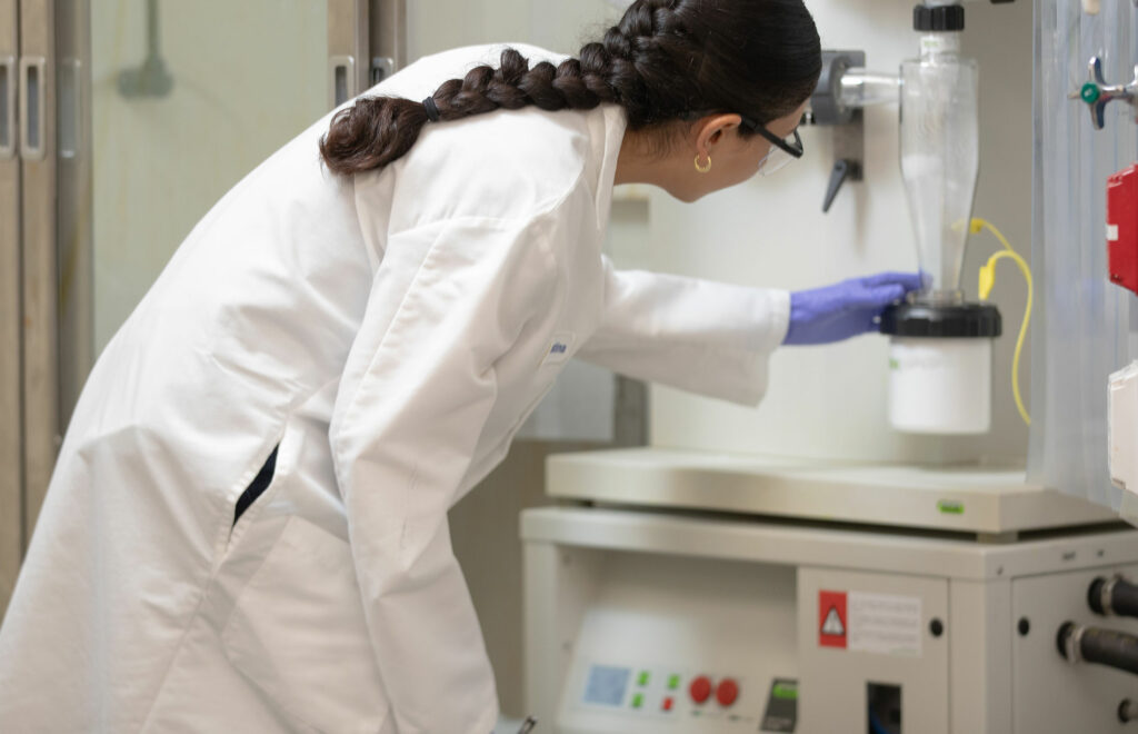 A woman in a lab coat and gloves operates laboratory equipment, adjusting a component in a lab setting.