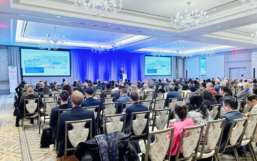 Conference attendees seated in a ballroom, facing a speaker on stage. Two screens display a presentation. The room is well-lit with chandeliers.
