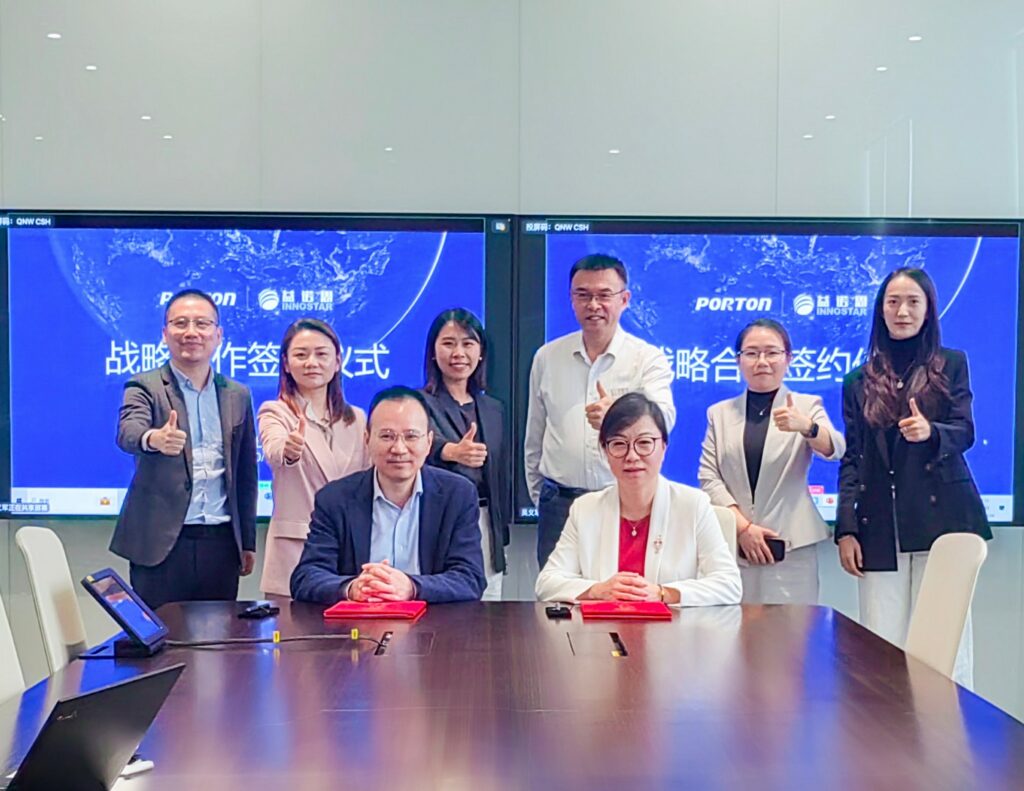 A group of eight people in business attire pose with thumbs up in a conference room, celebrating a strategic partnership. Two are seated at the table, while a screen behind them showcases business logos and text from Porton Pharma Solutions and Shanghai InnoStar.