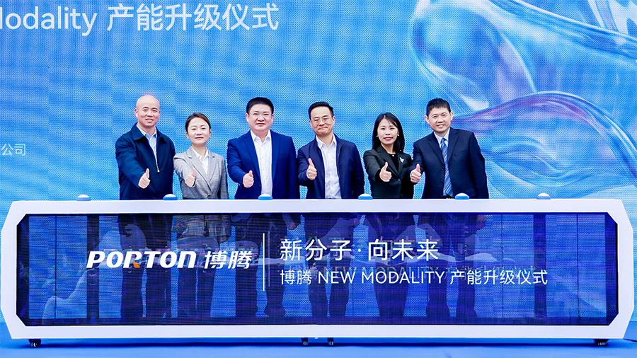 A group of six people in suits stands behind a display table with "PORTON" and Chinese text, giving thumbs up at a company event celebrating the modality capacity upgrade.